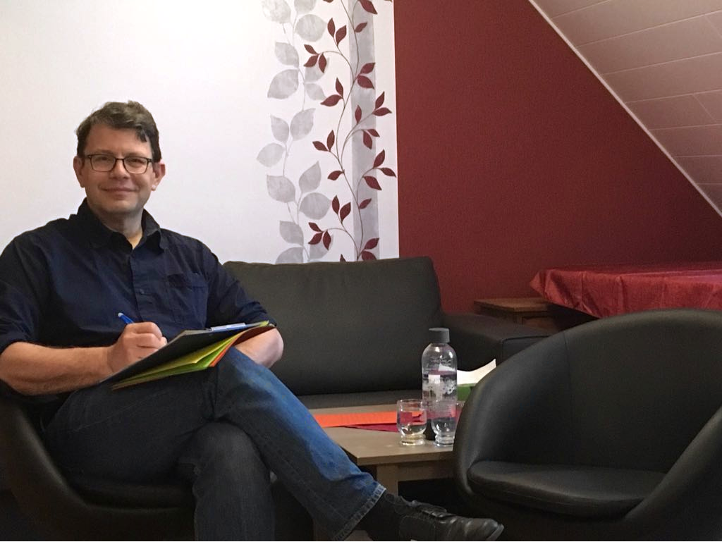 Manfred Kraut sitzt mit einem Schreibblock auf einem Drehsessel, gegenüber einem leeren Drehsessel